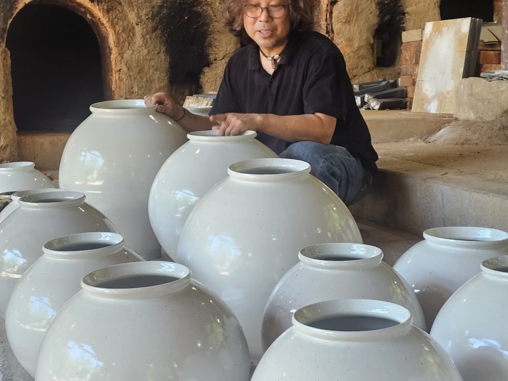 Shin Hyun Moon parle, accroupi derrière une rangée de moon jars blancs devant un four à bois